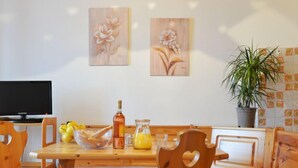 Ein Essbereich aus Holz mit Tisch, Stühlen und einer Bank. Ein Fernseher steht auf einem Schrank, es gibt einen Blumentopf und ein Bild an der Wand.