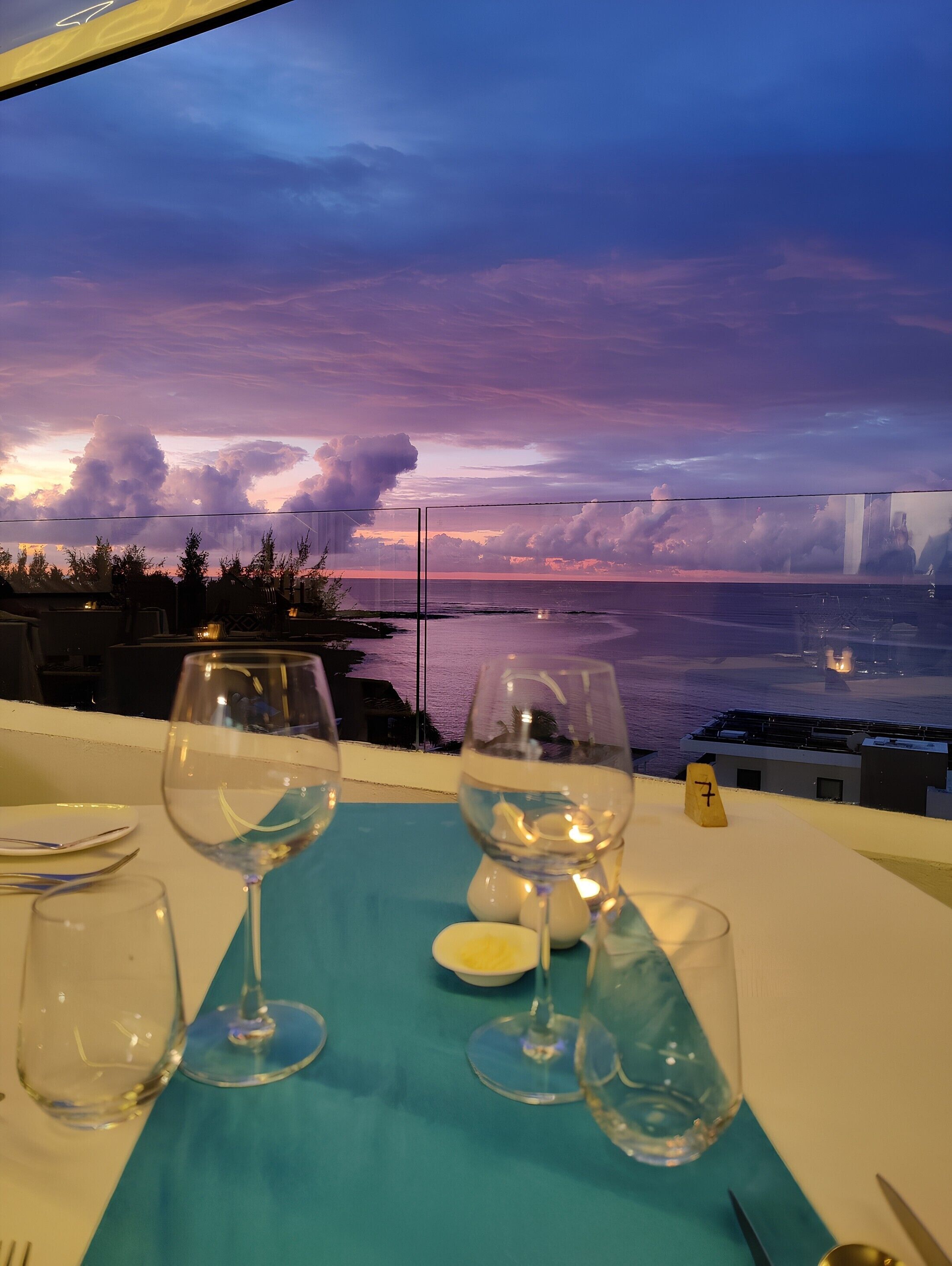 A table with empty wine glasses and a candle, overlooking a sunset view of the ocean and a bridge.
