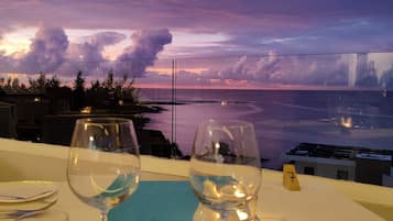 A table with empty wine glasses and a candle, overlooking a sunset view of the ocean and a bridge.