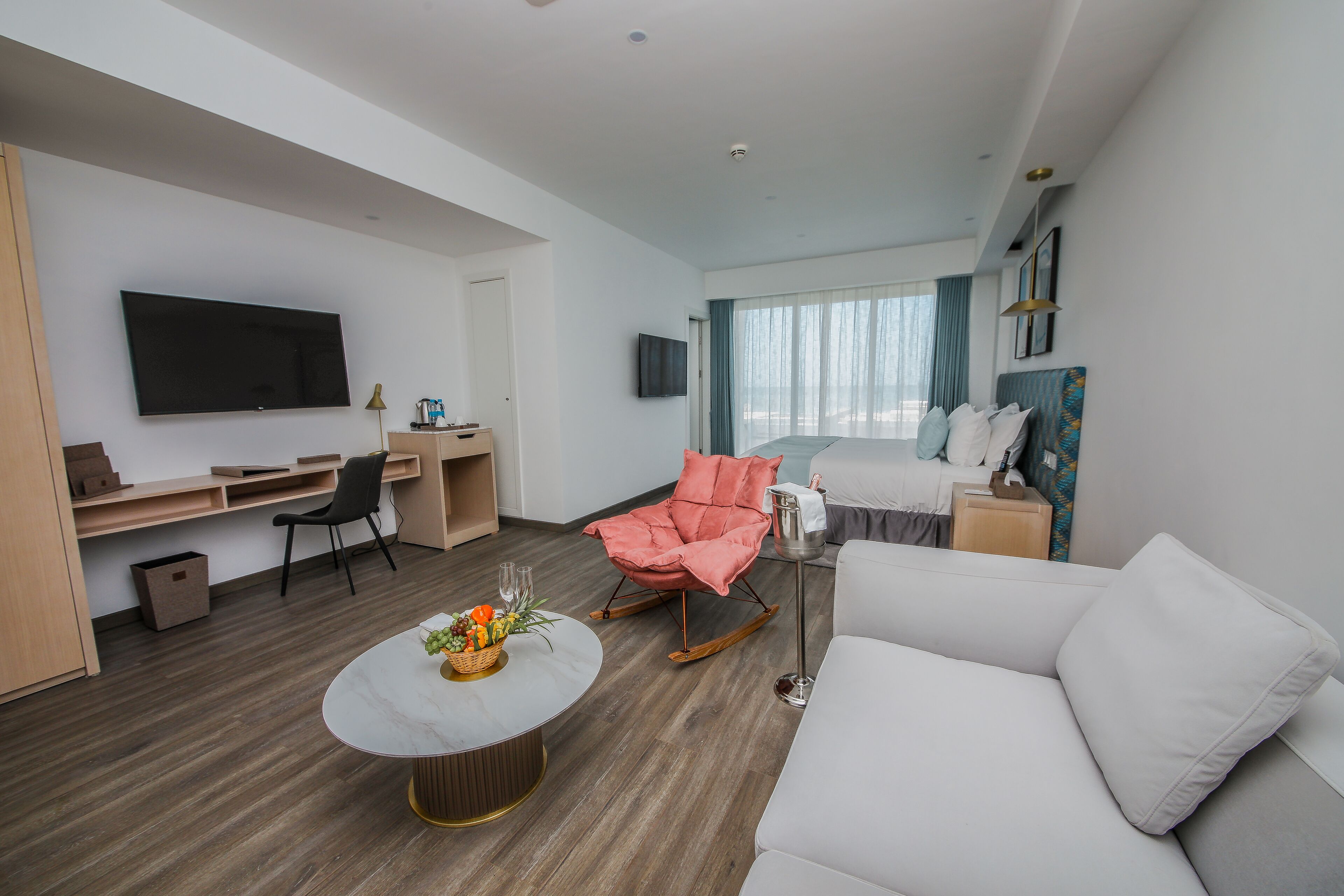 Une chambre d’hôtel moderne avec un canapé blanc, un fauteuil à bascule rose, un bureau avec un ordinateur, un téléviseur à écran plat et une grande fenêtre avec des rideaux.