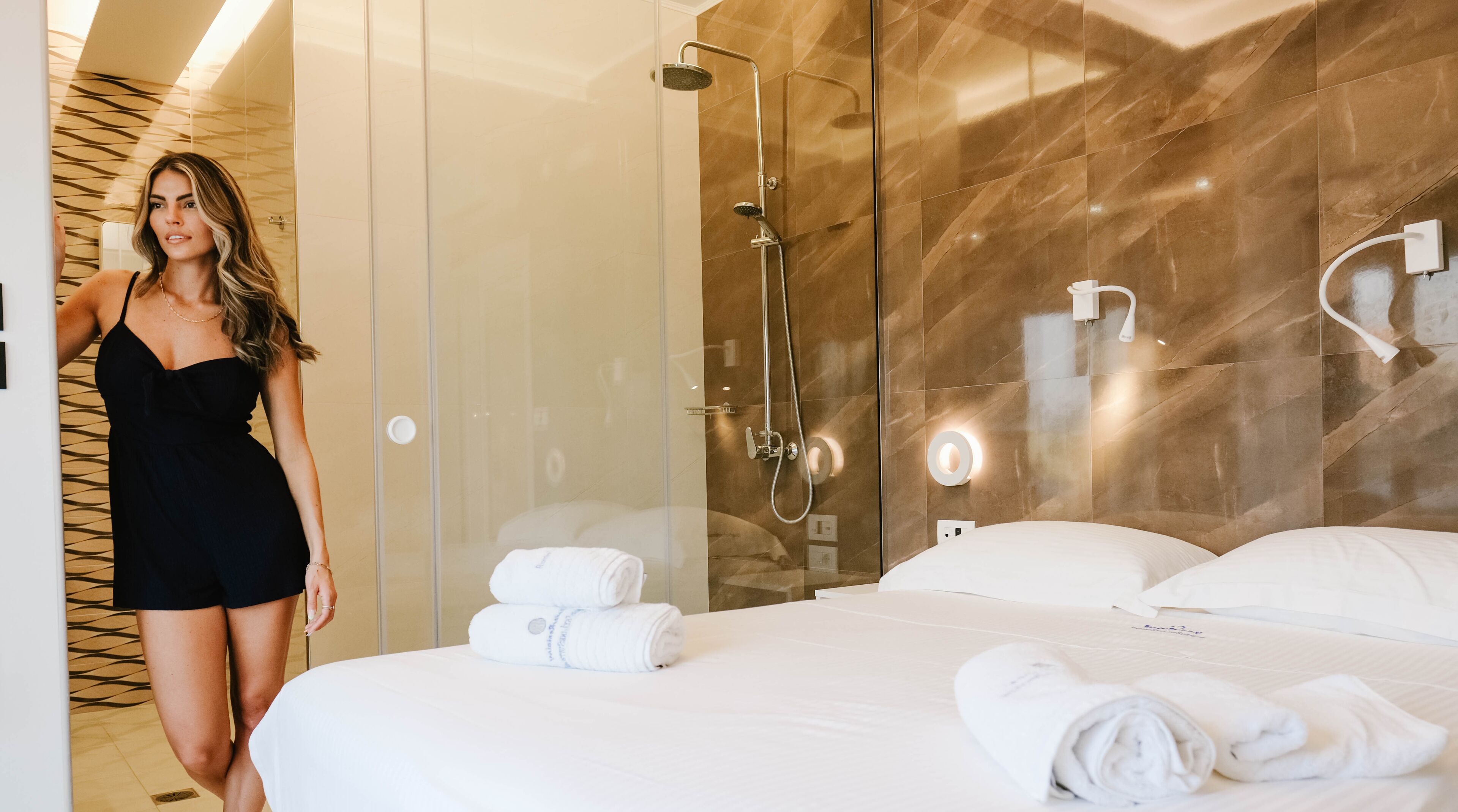 Moderni hotellihuone, jossa on lasiseinäinen suihkutila, siististi pedattu sänky ja näkyvillä oleva pyyheteline.