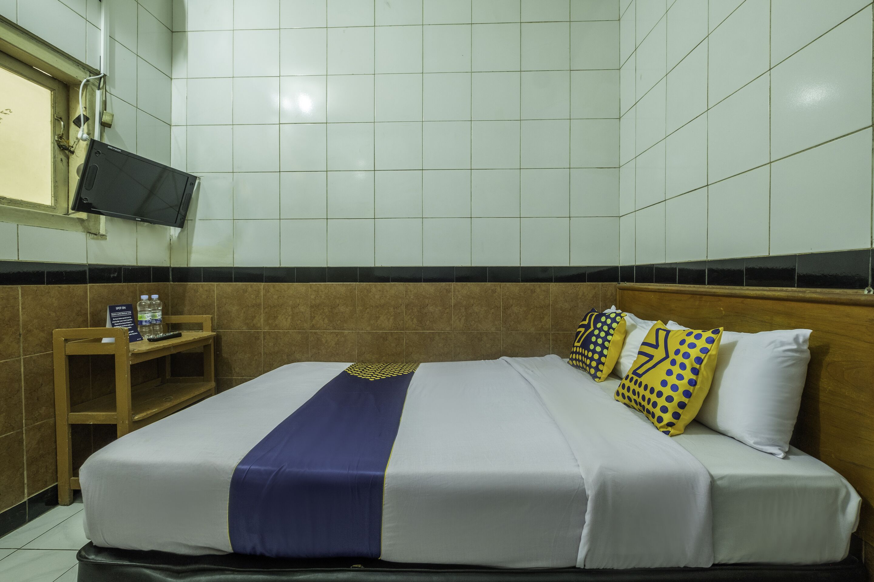A hotel room with a bed, a TV mounted on the wall, a small table with a water bottle, and a sign.