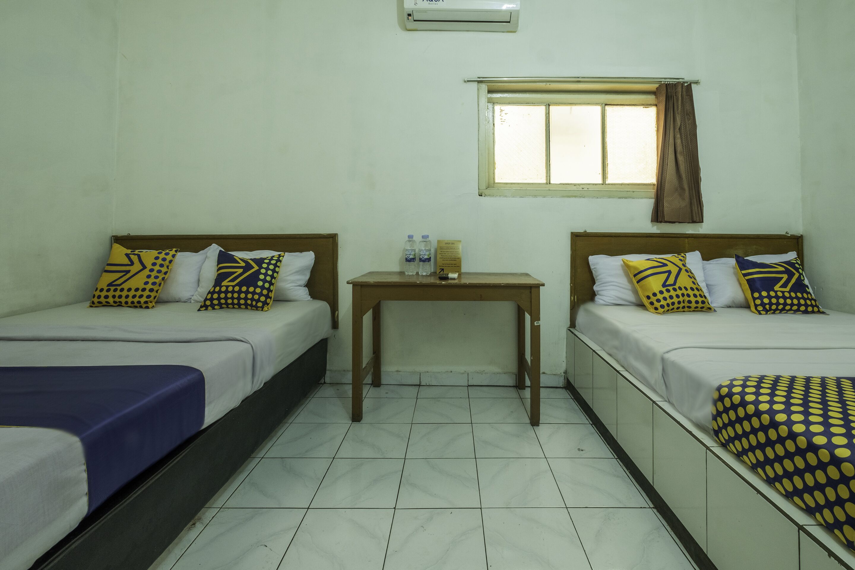 A room with two beds, each with patterned pillows and a blue blanket. There is a small table with two bottles and a box.
