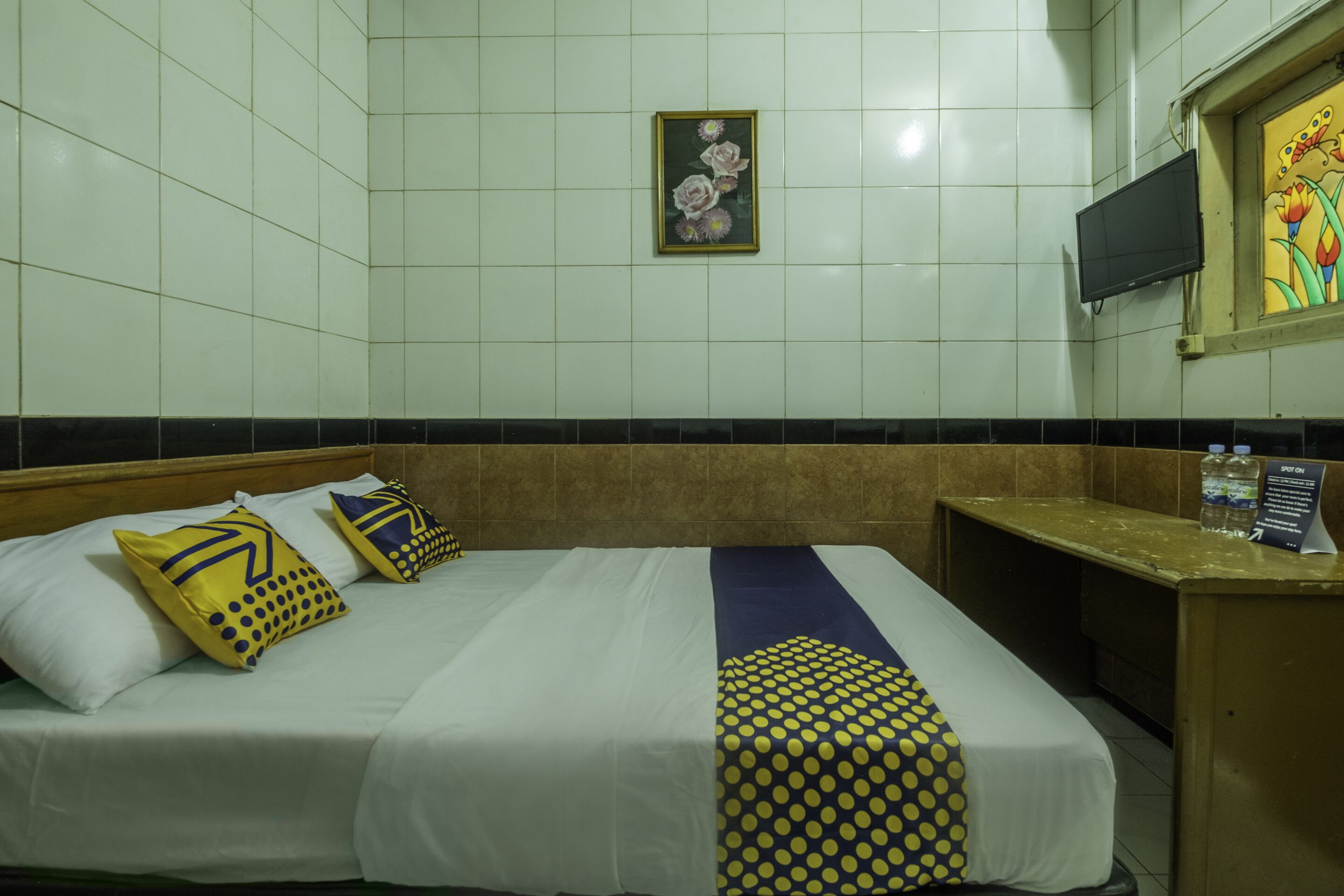 Una habitación de hotel con cama, almohadas, mesita de noche, televisor y un cuadro en la pared.