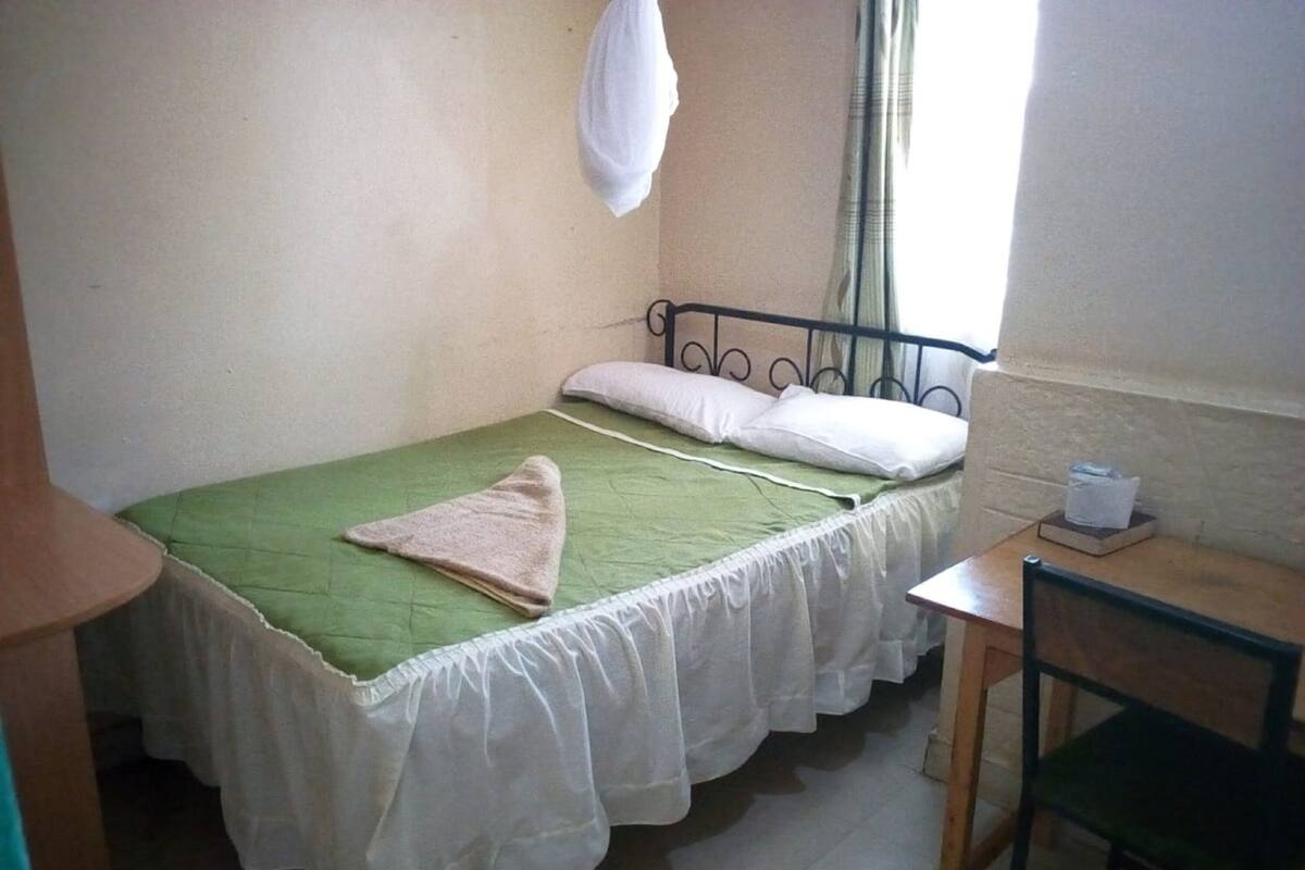 Ein kleines Zimmer mit einem Einzelbett, einer grünen Bettdecke, weißen Vorhängen, einem kleinen Tisch und einem Stuhl.