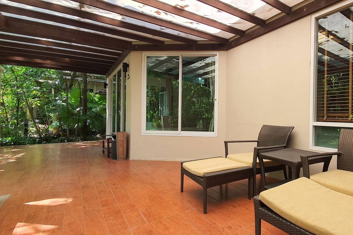 Un patio cubierto con mobiliario de mimbre y grandes ventanales con vistas a un jardín exuberante.