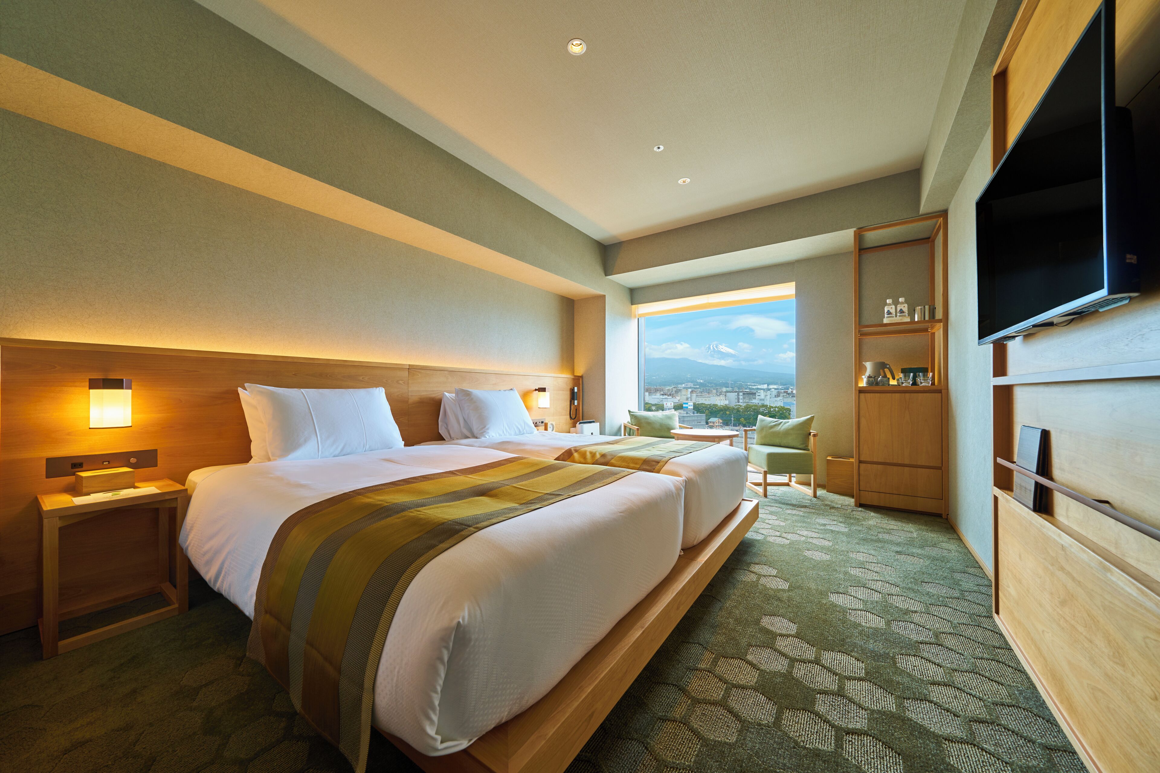 A hotel room with two beds, a TV, a bookshelf, and a view of a cityscape.