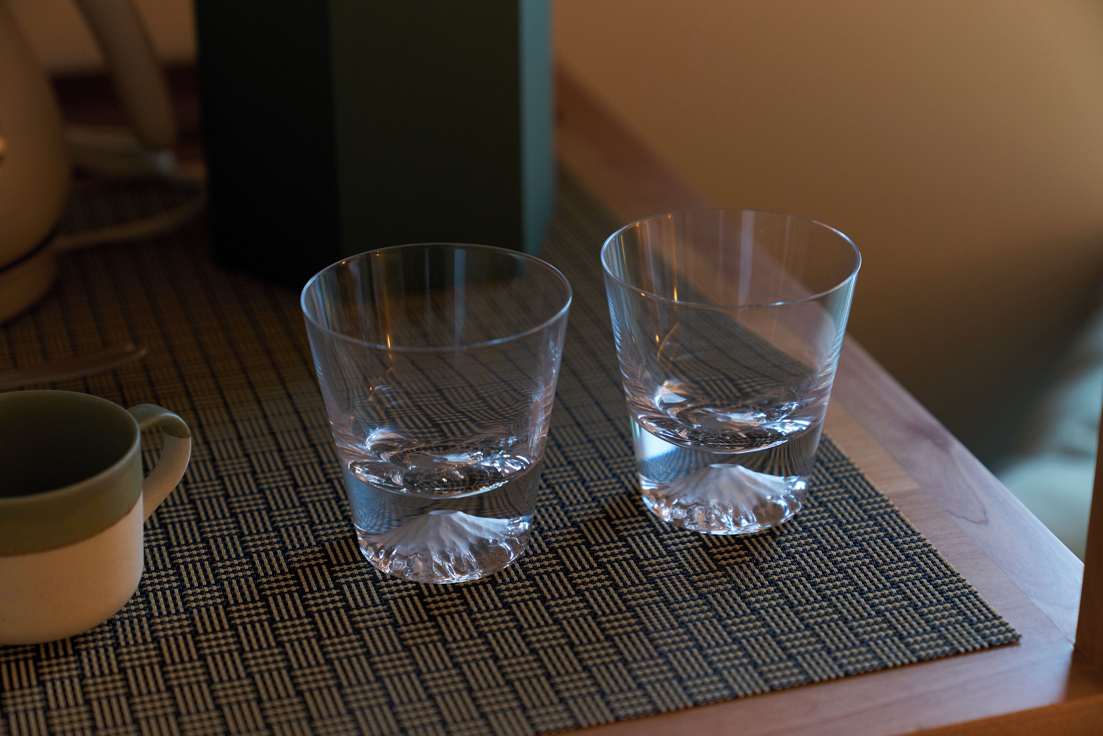 Two clear glasses of water on a woven placemat on a wooden table.
