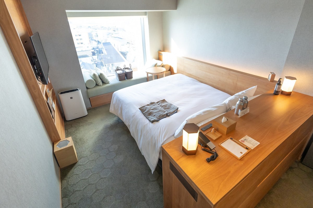 A hotel room with a bed, bedside table, lamp, and a window with a view of buildings.
