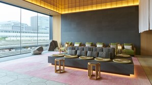 A modern lounge area with a sofa set, round wooden stools, and a large window offering a view of the cityscape. - Fujisan Mishima Tokyu Hotel (Mishima)