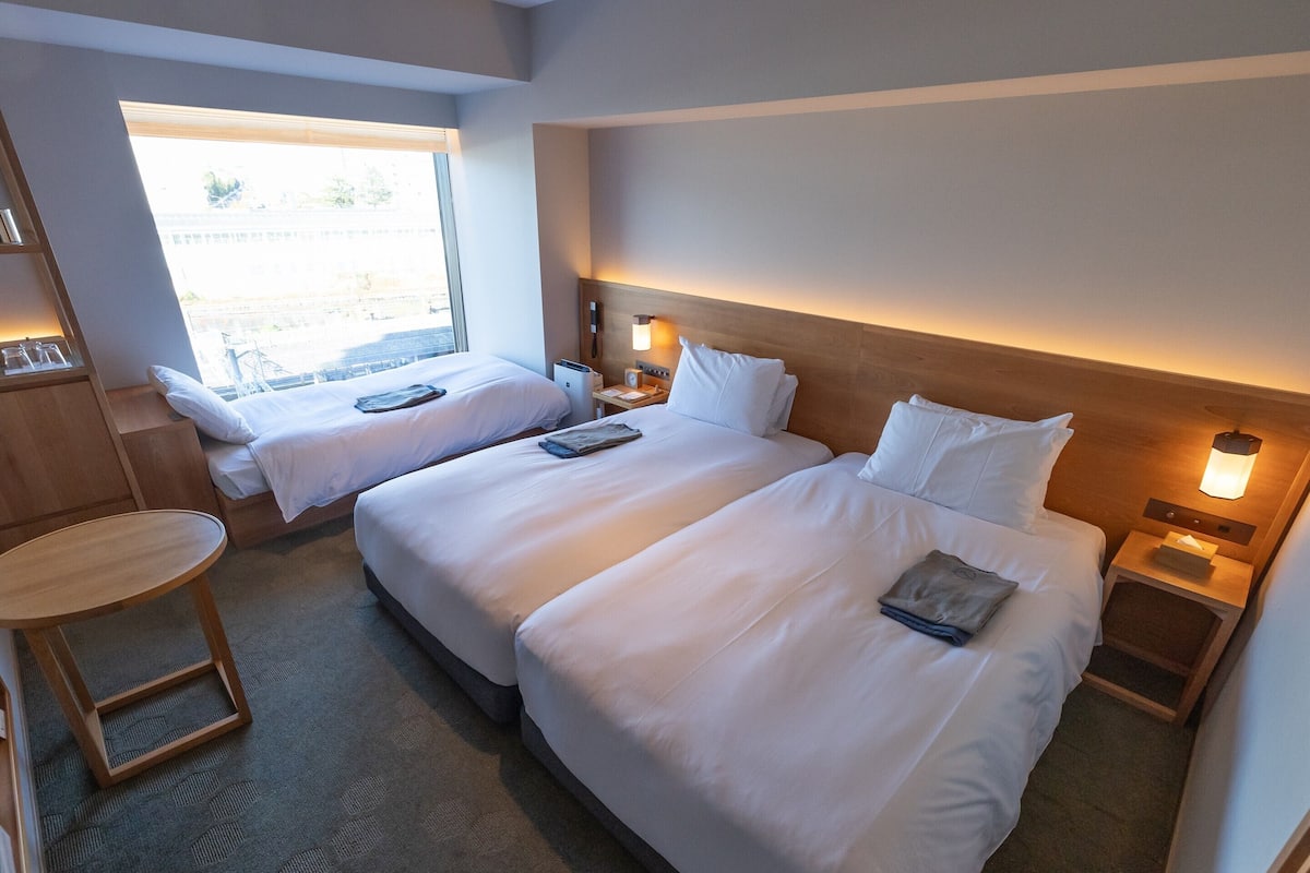 A hotel room with two beds, a small table, and a window with a view of the outside.