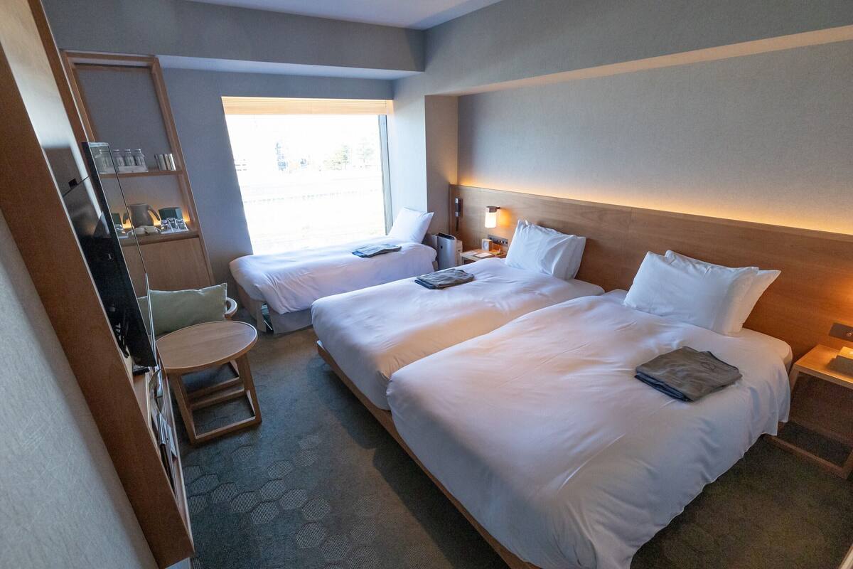 A hotel room with two beds, a small table, a chair, a mirror, and a window with a view of the outside.