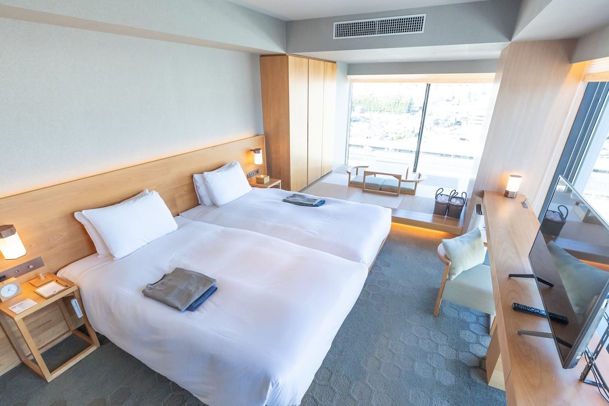 A hotel room with two beds, a desk, a chair, a TV, and a window with a view of the outside.