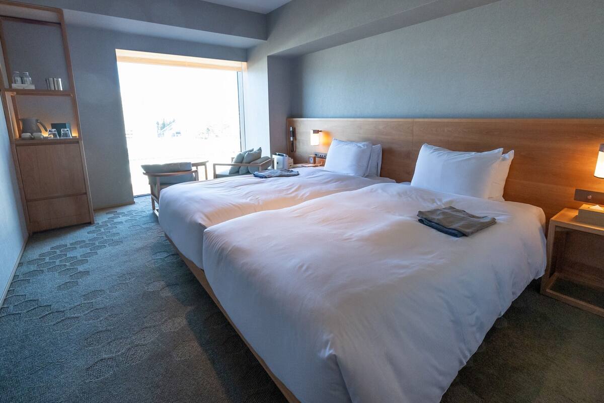 A hotel room with two beds, a wooden headboard, a bedside table with a lamp, a small desk with a chair, and a large window.