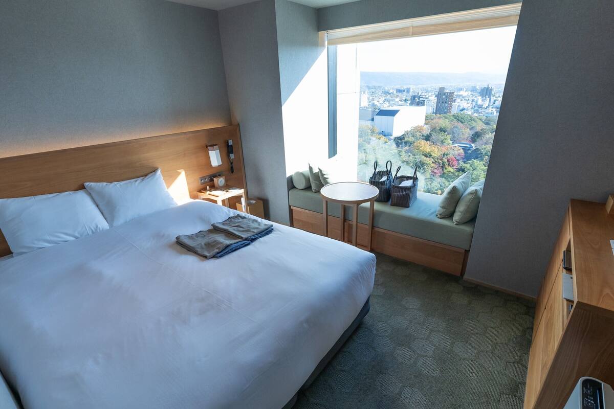 A hotel room with a large bed, a small table, a sofa with a view, and a desk with a lamp.