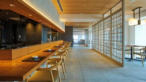 A modern restaurant with a long counter, wooden chairs, and a view of the city through large windows. - Fujisan Mishima Tokyu Hotel (Mishima)