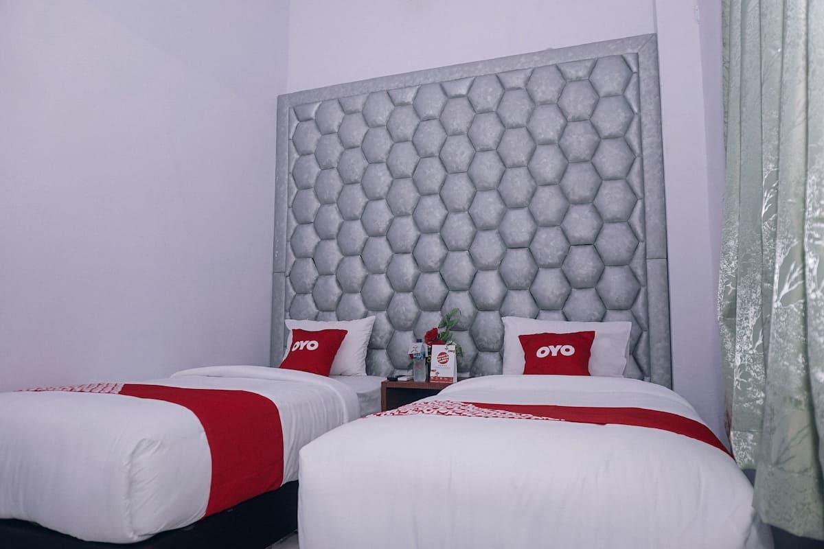 A hotel room with two beds, each with white bedding and red accents, a grey textured headboard, and a small table with a vase and a box.
