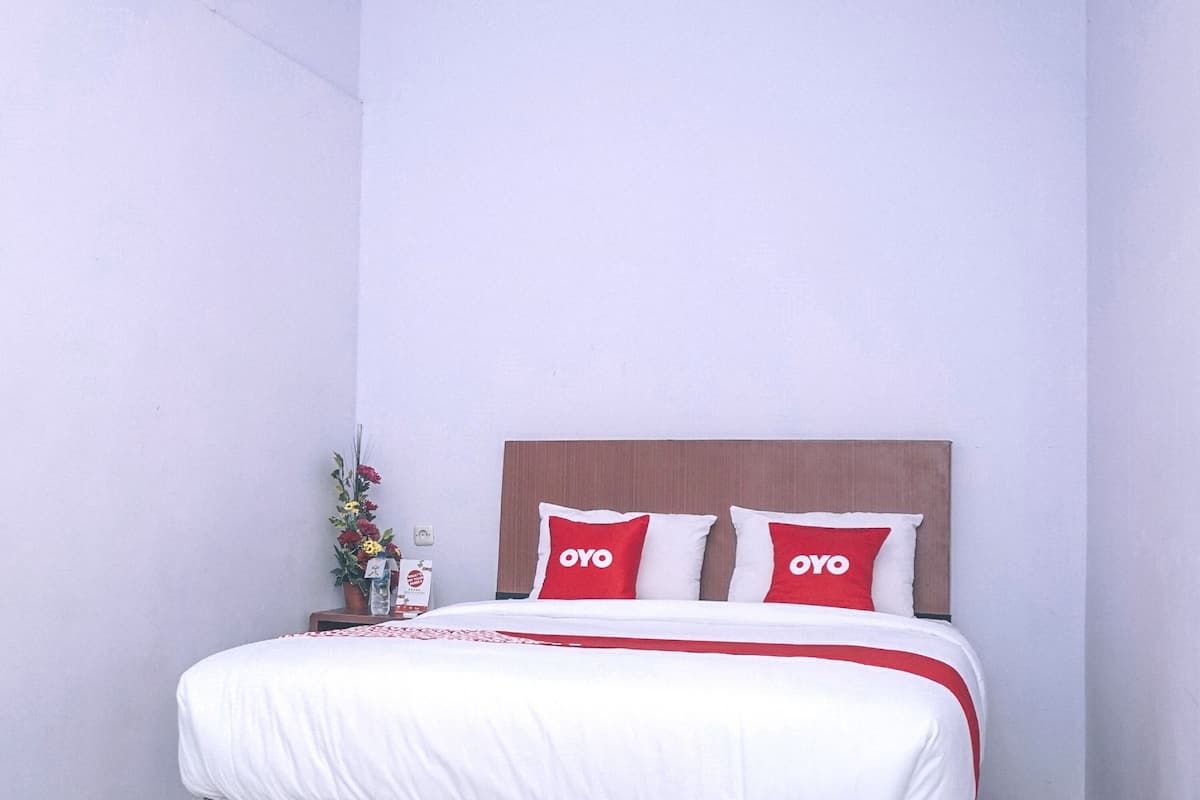 A neatly made bed with white bedding and red pillows, a wooden headboard, and a small bedside table with a vase of flowers.