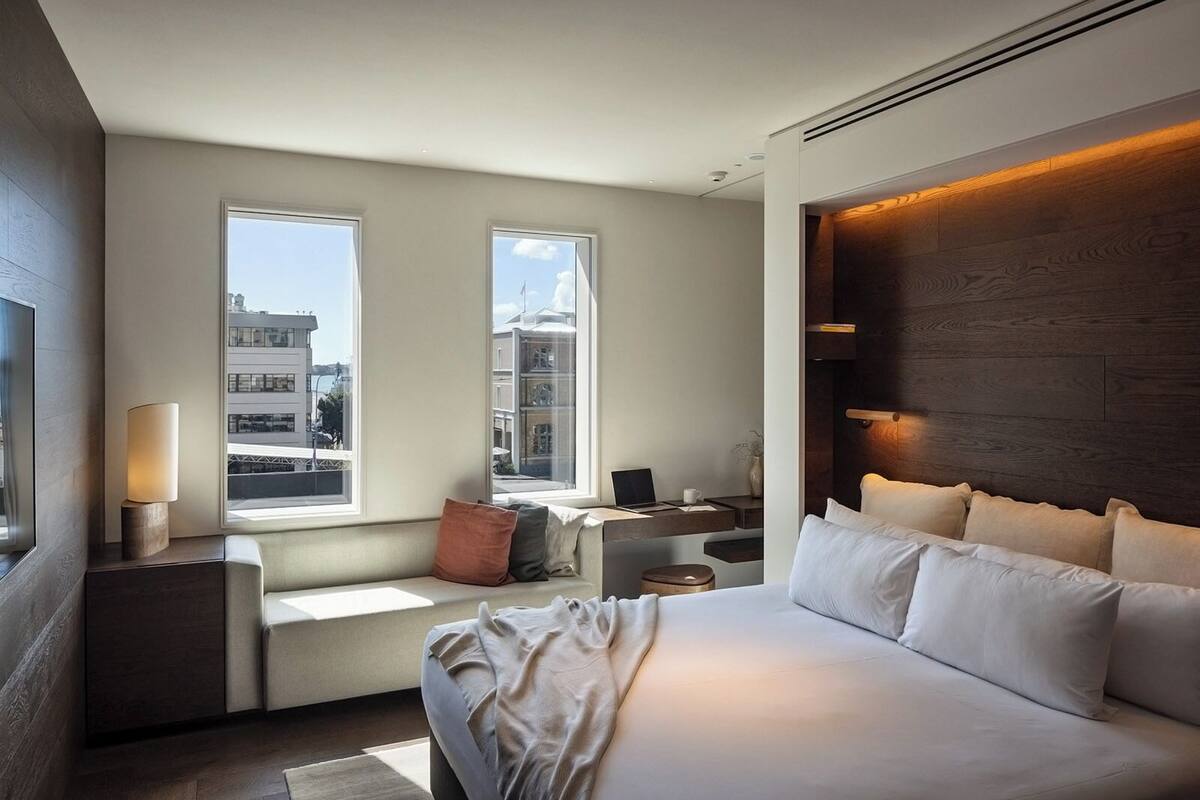 A modern hotel room with a large bed, a desk with a laptop, and two windows offering a city view.