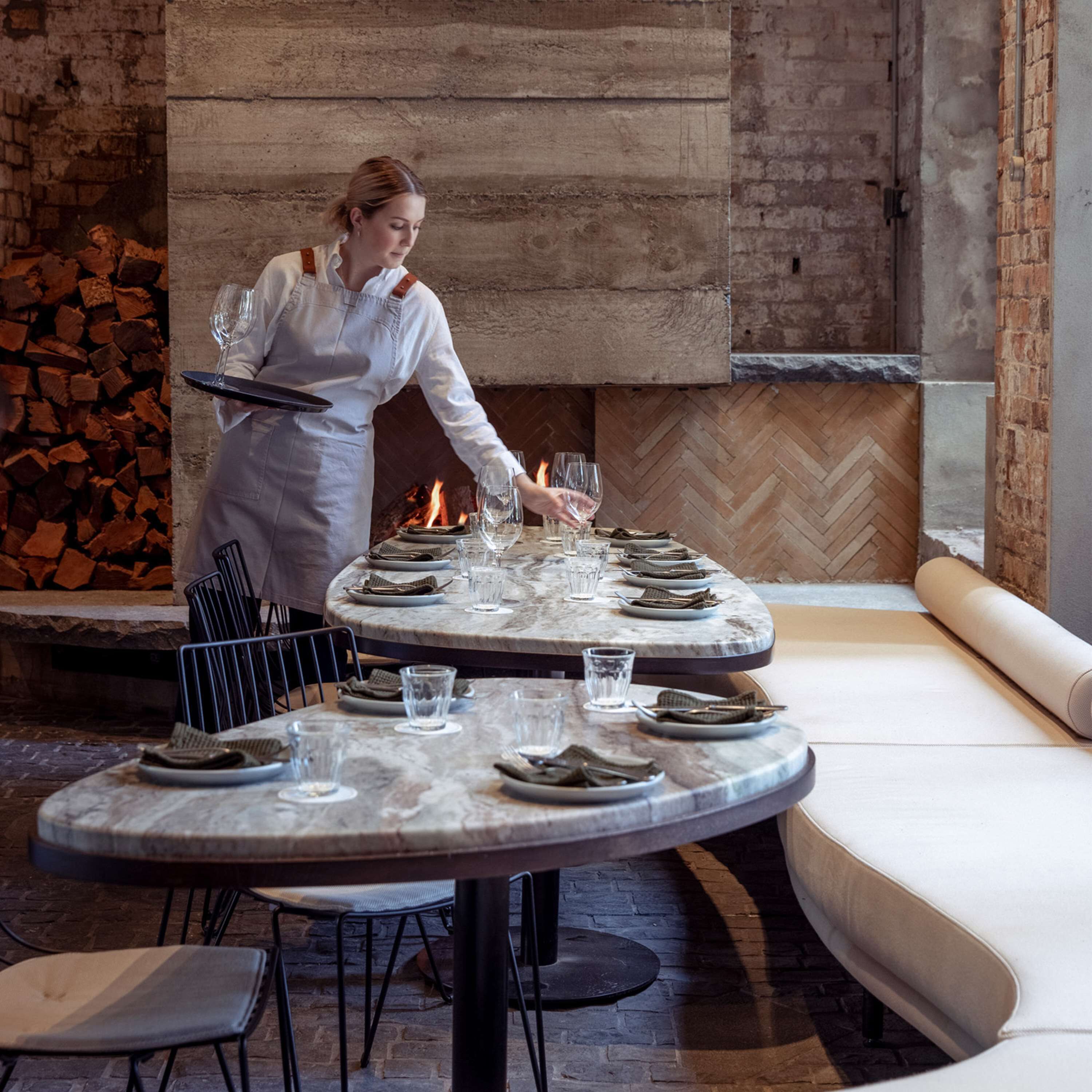 A restaurant with a round table set for a meal, a fire in the background, and a brick wall.