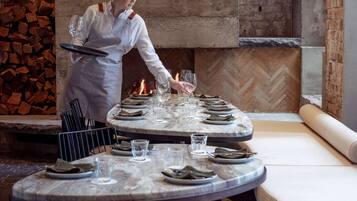 A restaurant with a round table set for a meal, a fire in the background, and a brick wall.