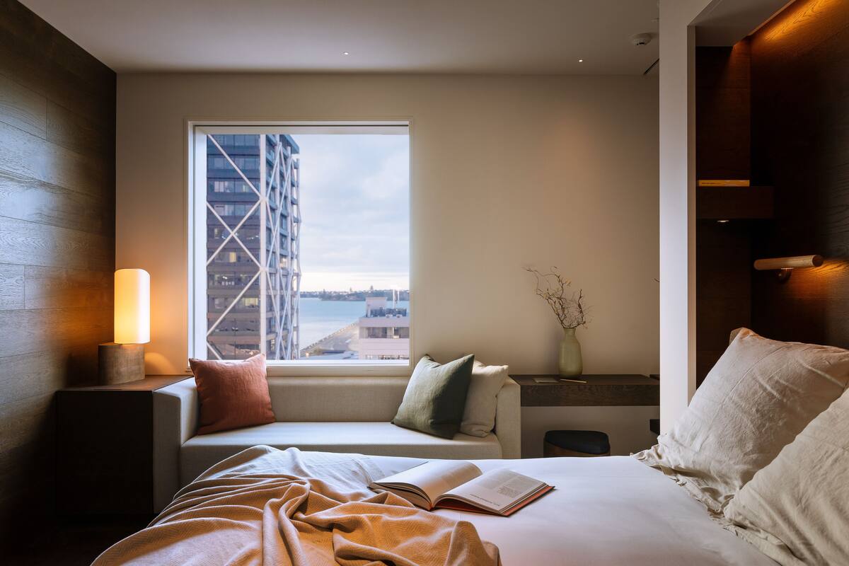 A modern hotel room with a large bed, a bedside table with a lamp, a window with a view of a building, and a small seating area with cushions.