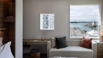 Habitació d'hotel moderna amb un gran finestral que ofereix vistes al port, un llit, una escriptori i una zona de seients amb coixins.