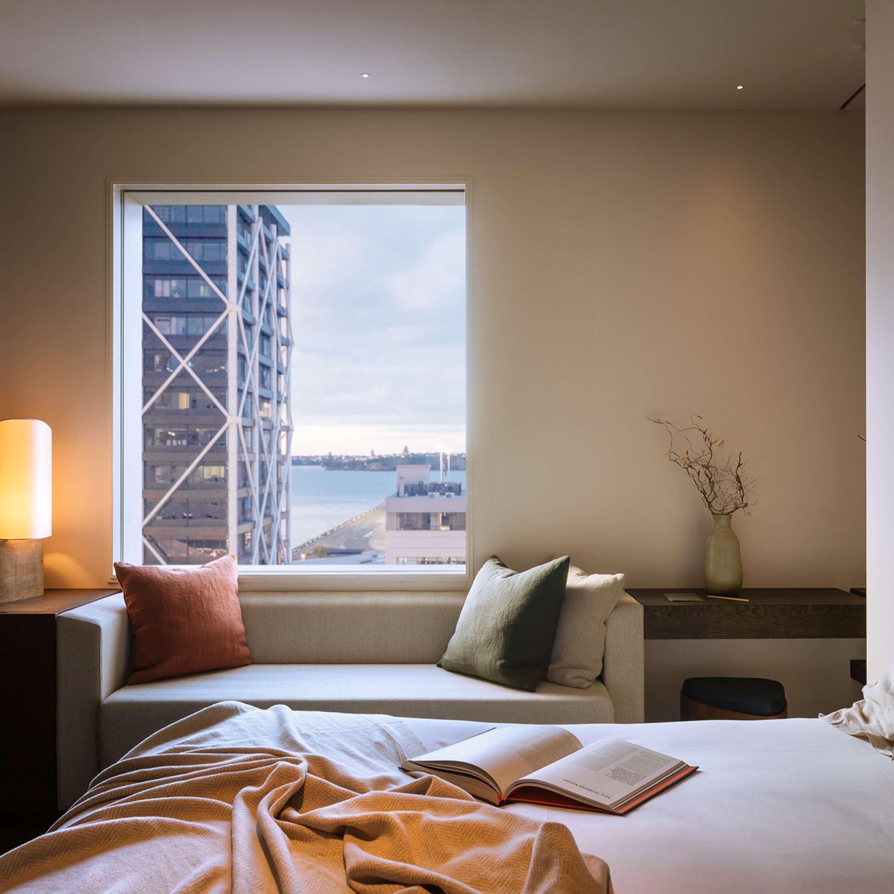 A modern hotel room with a large window, a sofa with cushions, a bedside table with a lamp, and a vase with branches.