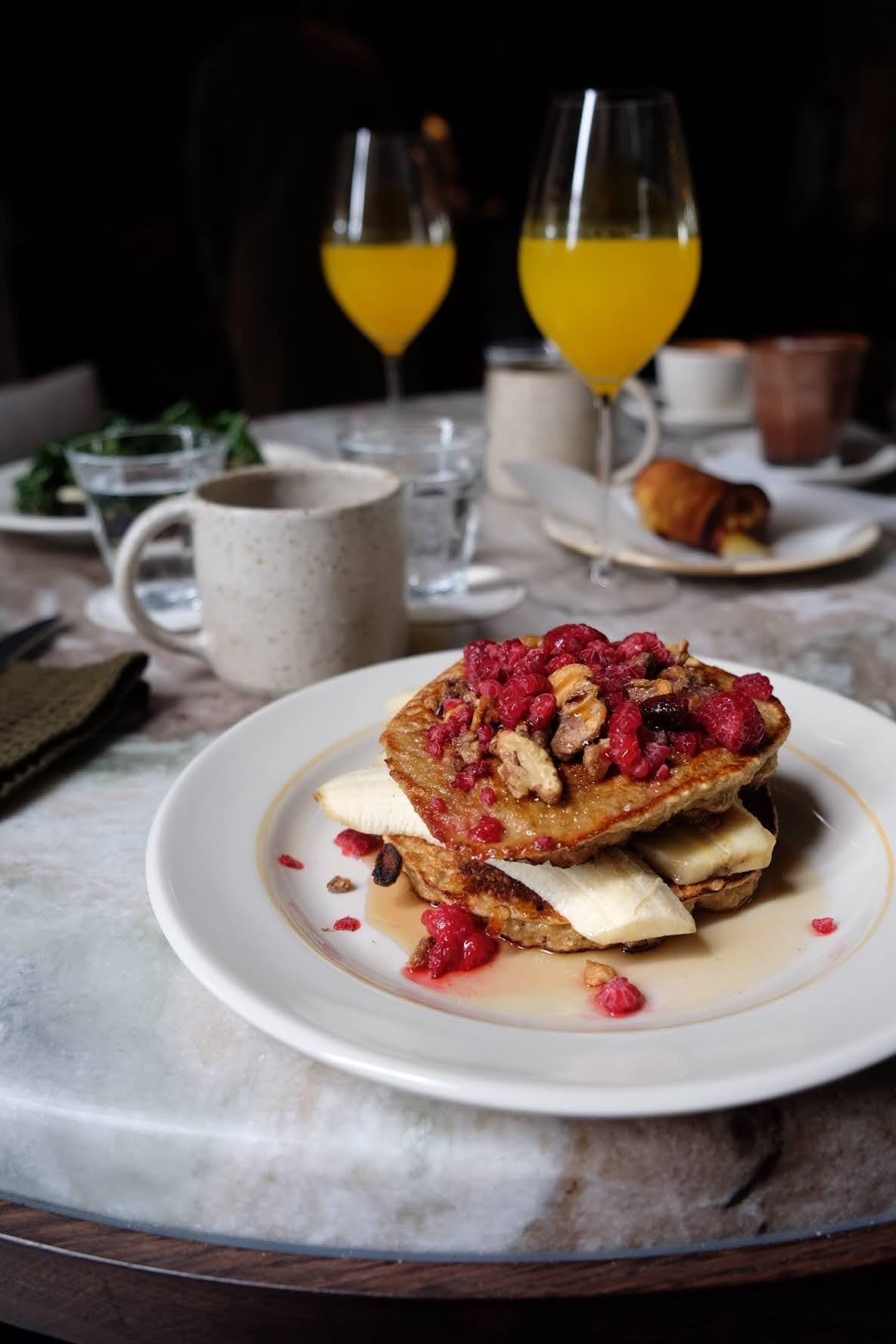 Un desayuno con una fuente de panqueques cubiertos de frutos rojos y nueces, una taza de café y dos vasos de jugo de naranja.