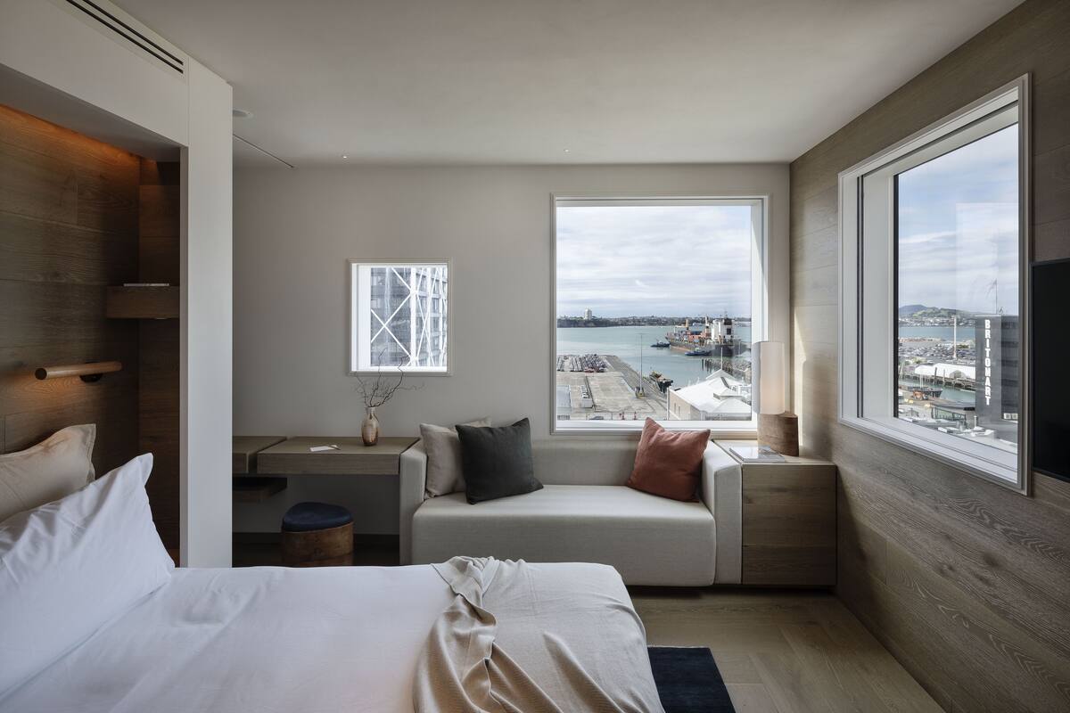 A modern hotel room with a large window offering a view of a harbor, a bed with white linens, a small seating area with cushions, and a wooden accent wall.