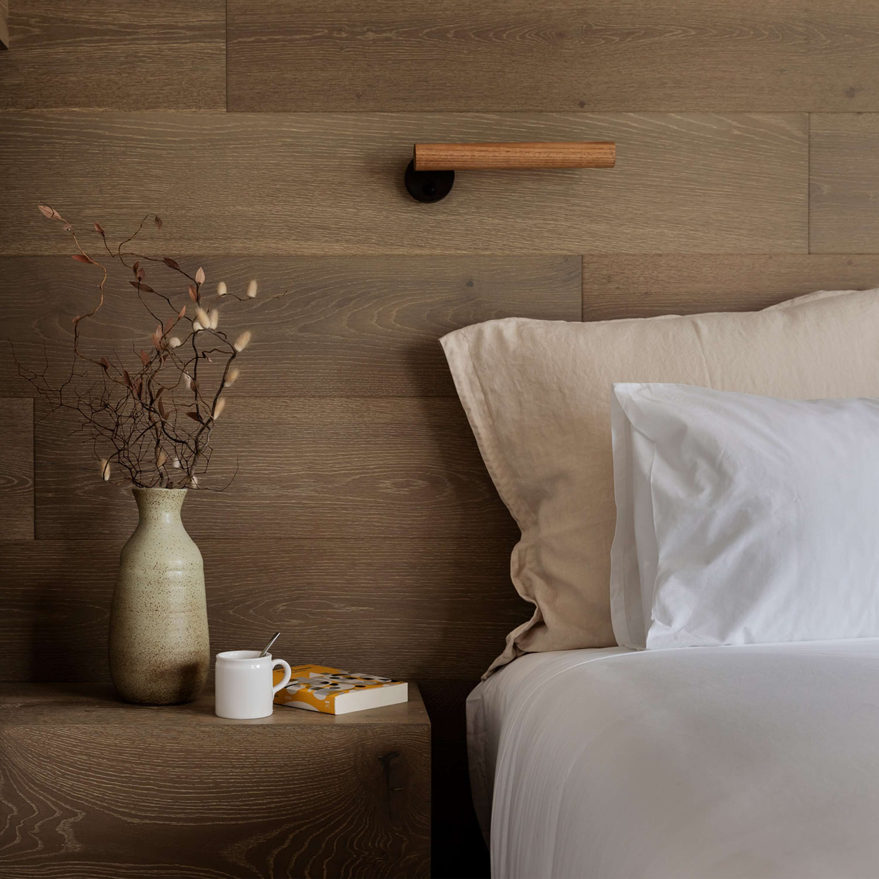 Una camera da letto con una testiera in legno, un comodino con un vaso di rami secchi, una tazza bianca e un libro.