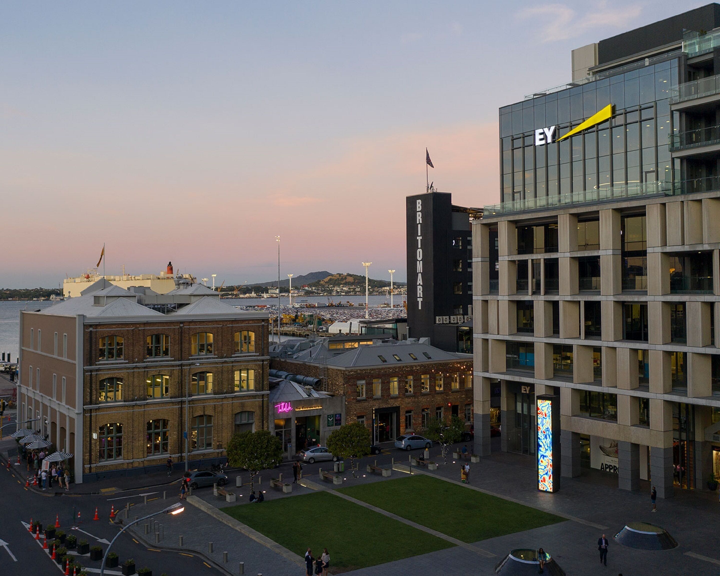 Un paysage urbain où se côtoient une architecture moderne et traditionnelle, avec notamment un bâtiment imposant arborant l’enseigne « EY » et une tour « Britomart ».