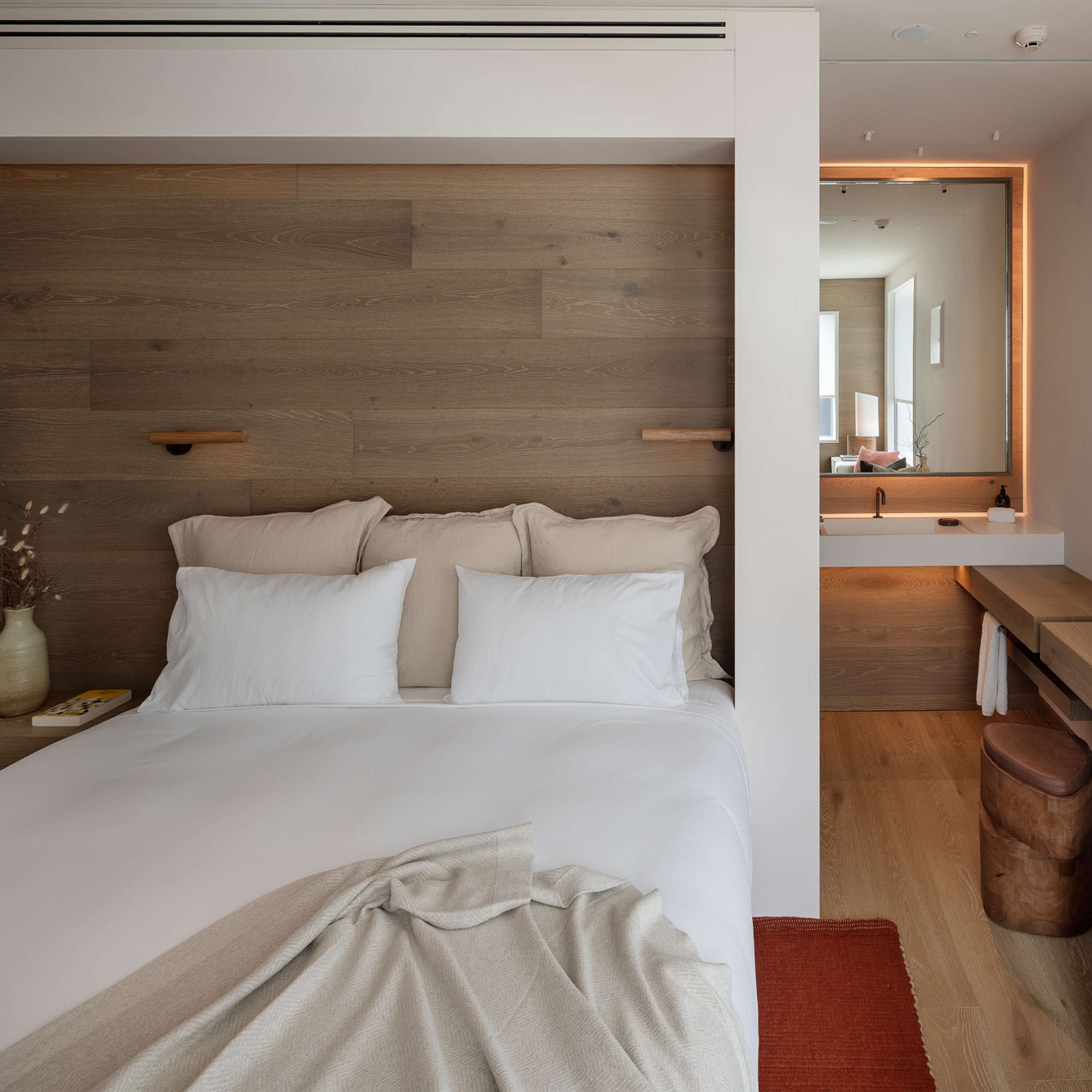 Une chambre moderne avec une tête de lit en bois, un grand lit avec du linge de lit blanc, une table de chevet avec un livre, et une salle de bain avec un miroir et un lavabo.
