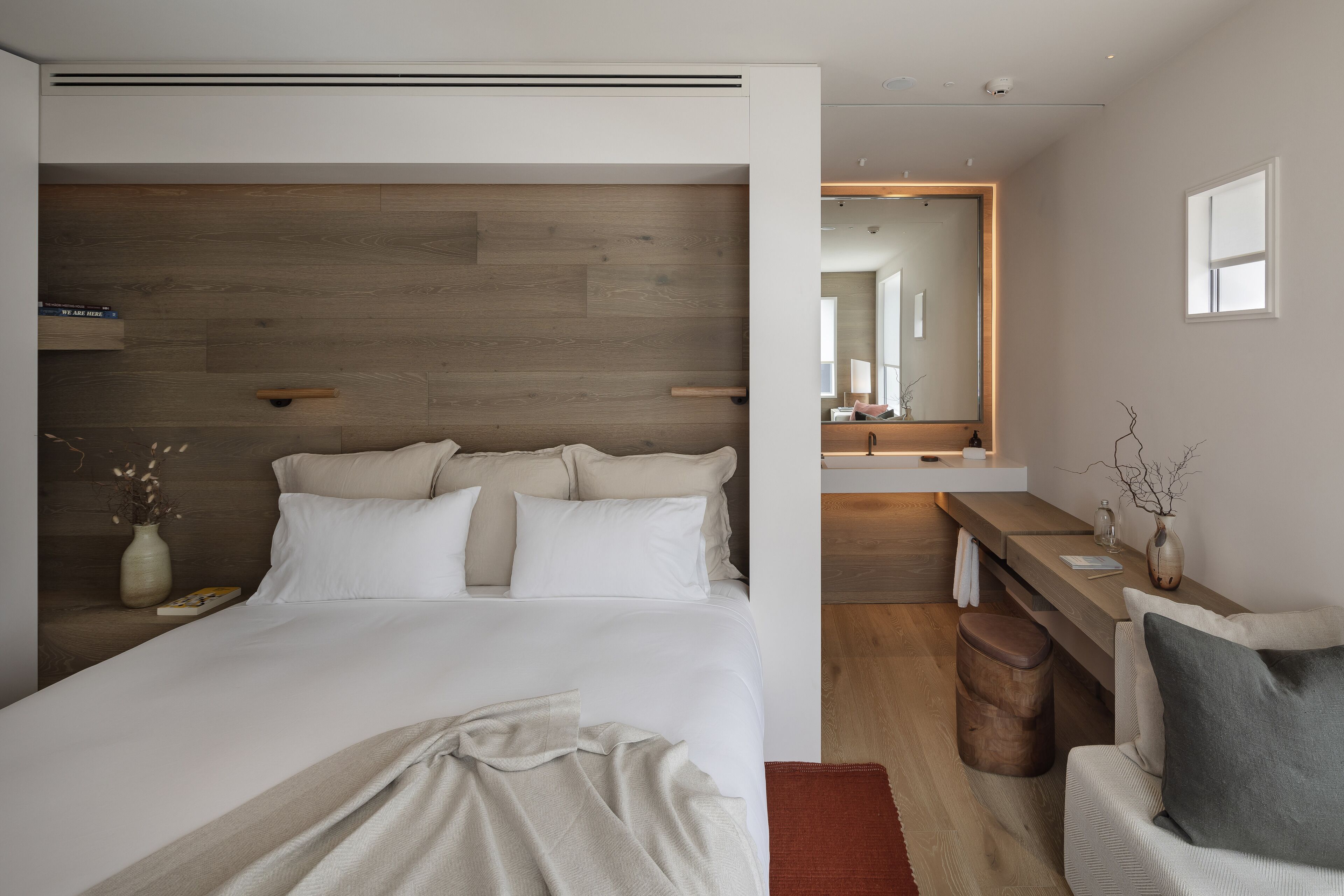 Un dormitorio moderno con una cama grande, un cabecero de madera, una mesita de noche con un florero y un banco con un florero.