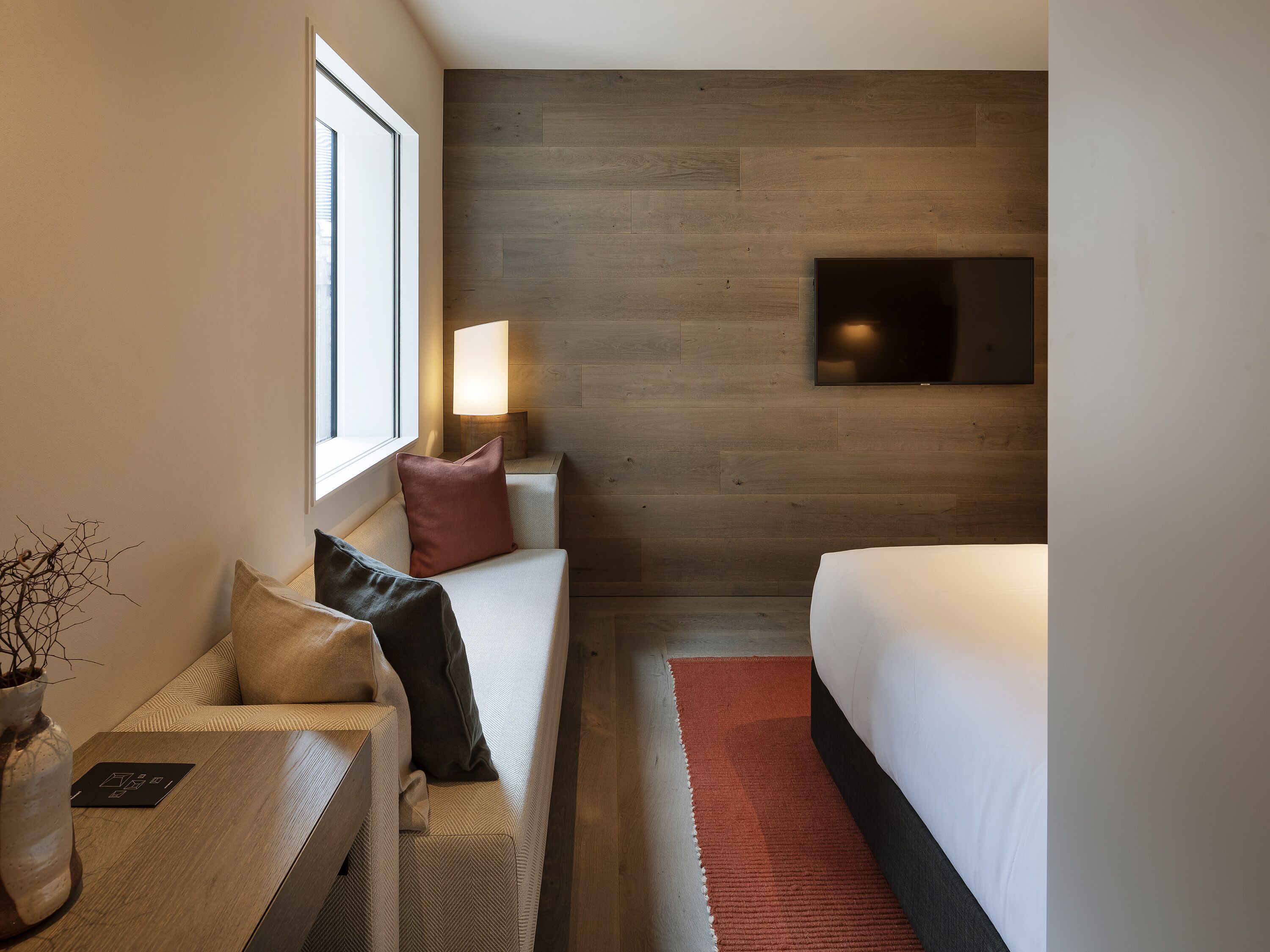 Quarto de hotel moderno com uma janela grande, uma televisão de tela plana na parede, um sofá com almofadas, uma mesa de cabeceira e um tapete vermelho.