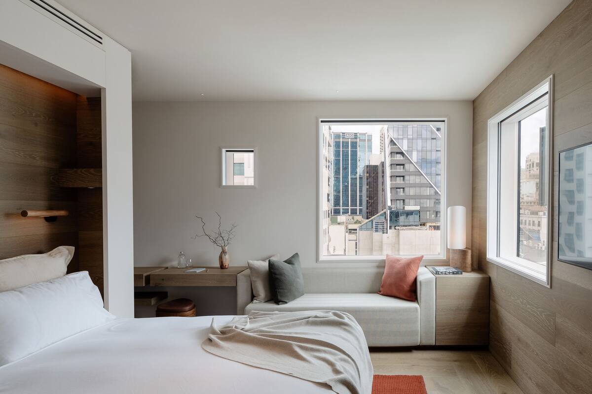 A modern hotel room with a large bed, a desk with a vase, a window with a city view, and a small seating area.