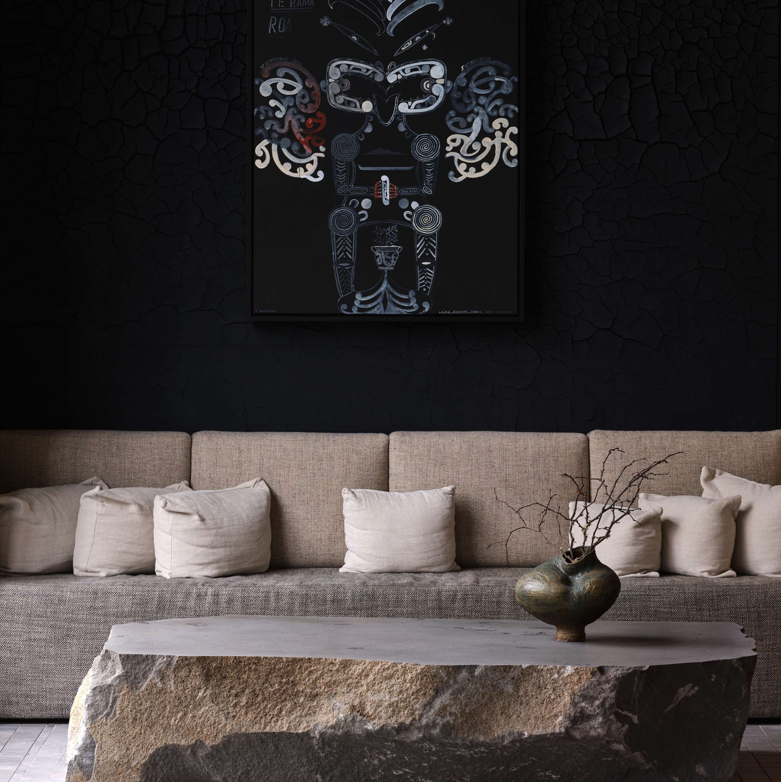 Ein modernes Wohnzimmer mit dunkler Wand, einer Couch mit vielen Kissen und einer dekorativen Vase auf einem Steintisch.