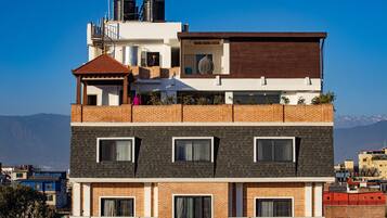 Un edificio de varios pisos con terraza en la azotea, balcón cubierto y vistas a montañas distantes.