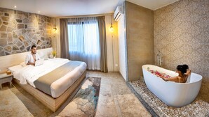 A modern hotel room with a stone wall, a large bed, a sitting area, and a bathtub with rose petals. - Arushi Boutique Hotel (Kathmandu)