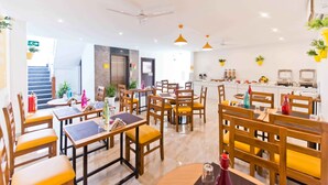 A modern dining area with wooden tables and chairs, a kitchen in the background, and a ceiling fan. - X by Bloom - Hebbal (Bengaluru)