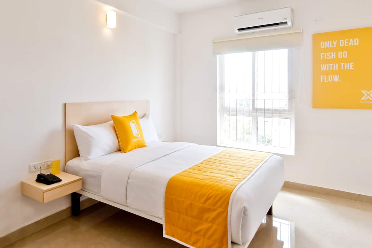 Quarto de hotel com cama, mesa de cabeceira com telefone, janela e uma frase motivacional na parede.