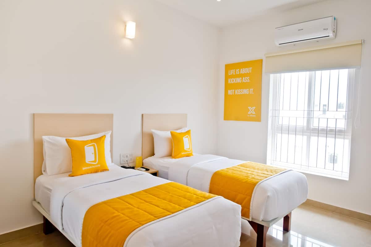 Quarto de hotel com duas camas, cada uma com colcha branca e amarela, uma cama simples com travesseiro branco e amarelo, uma mesa de cabeceira, um aparelho de ar condicionado e uma janela com vista para a cidade.