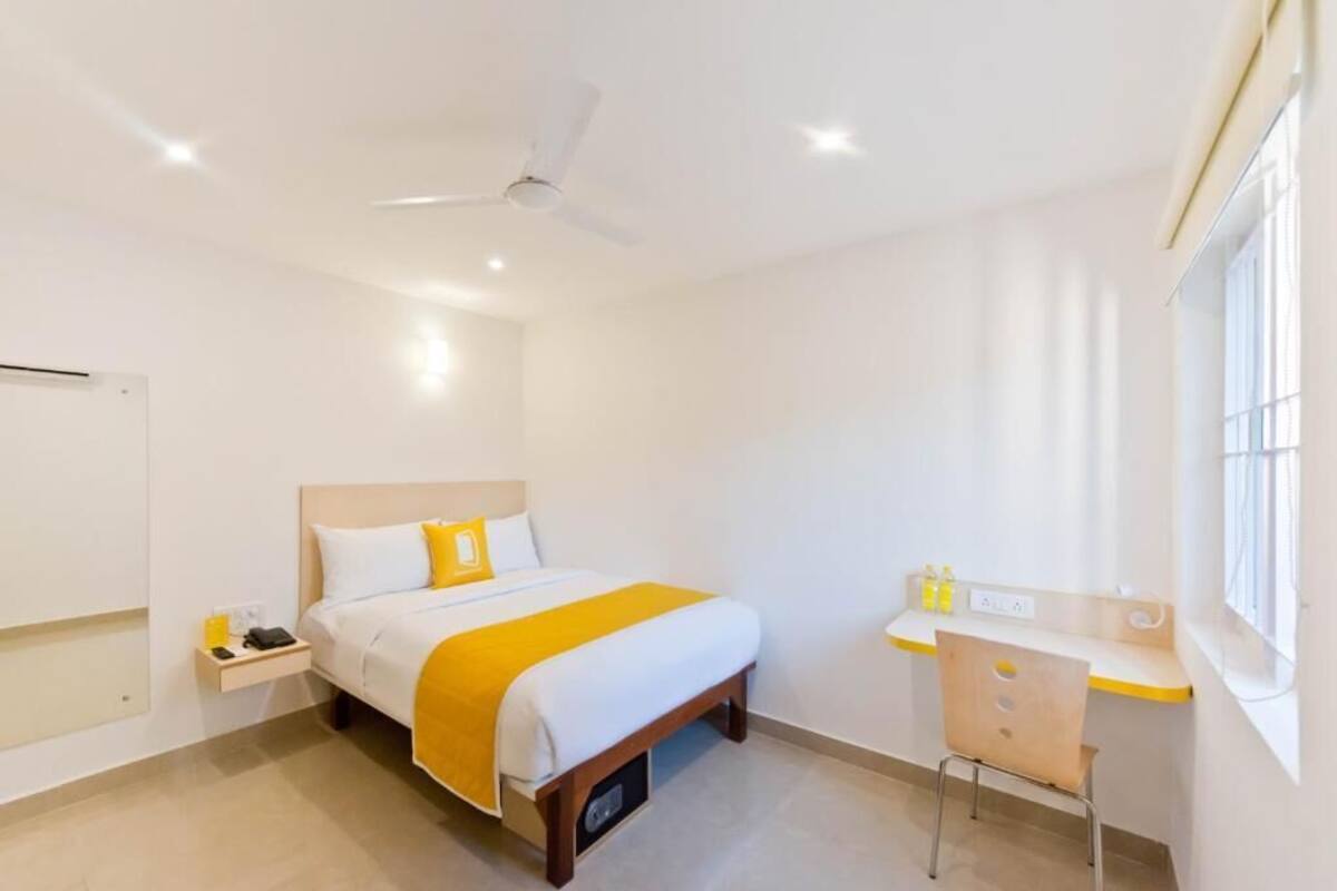 Quarto de hotel com cama, mesa de cabeceira, escrivaninha, cadeira e ventoinha no teto.