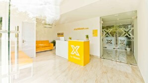 A reception area with a yellow desk, a white sofa, and a glass door entrance. - X by Bloom - Hebbal (Bengaluru)