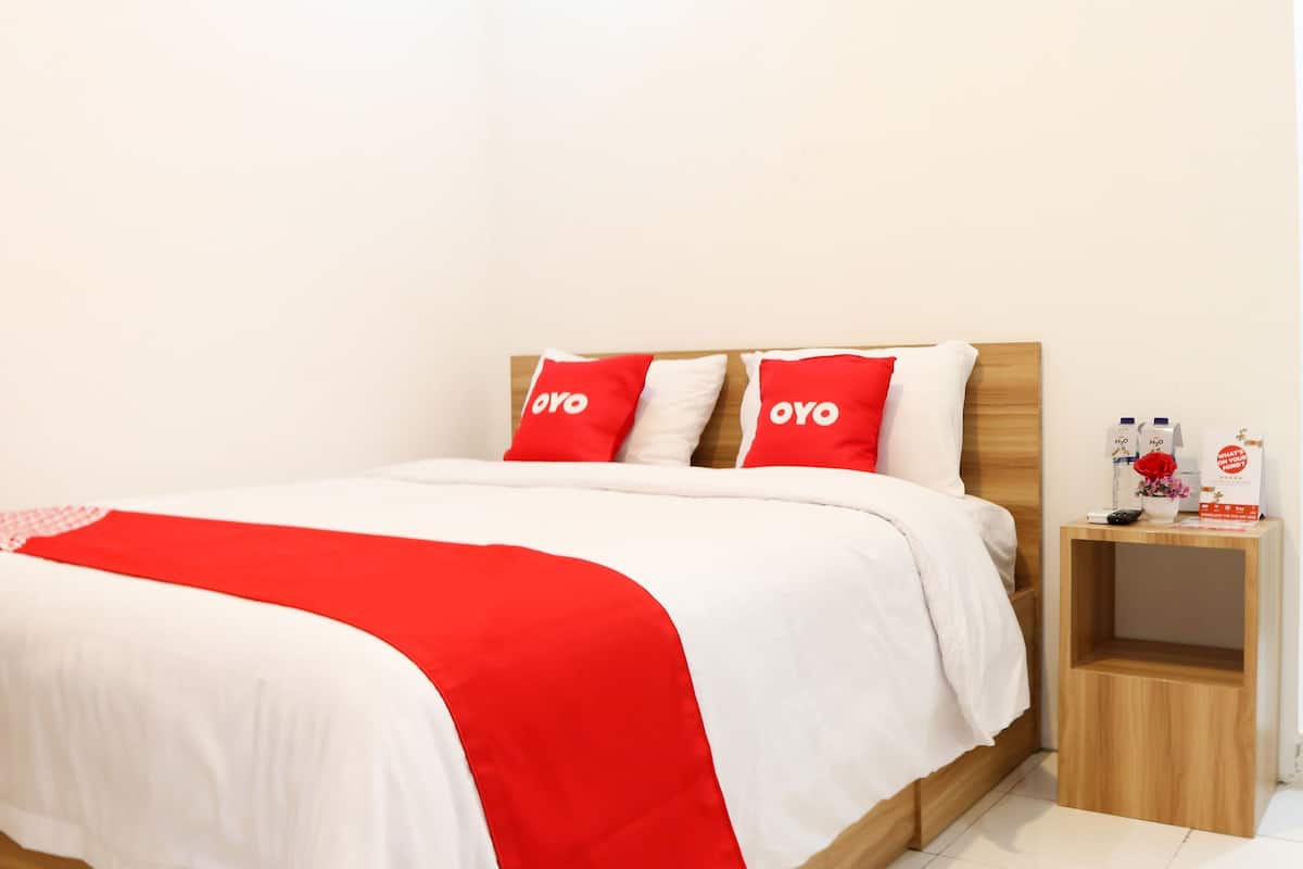 A neatly made bed with a white and red bedspread, two red pillows with the word "OYO" on them, and a wooden nightstand with a remote control, a bottle of water, and a glass of milk.