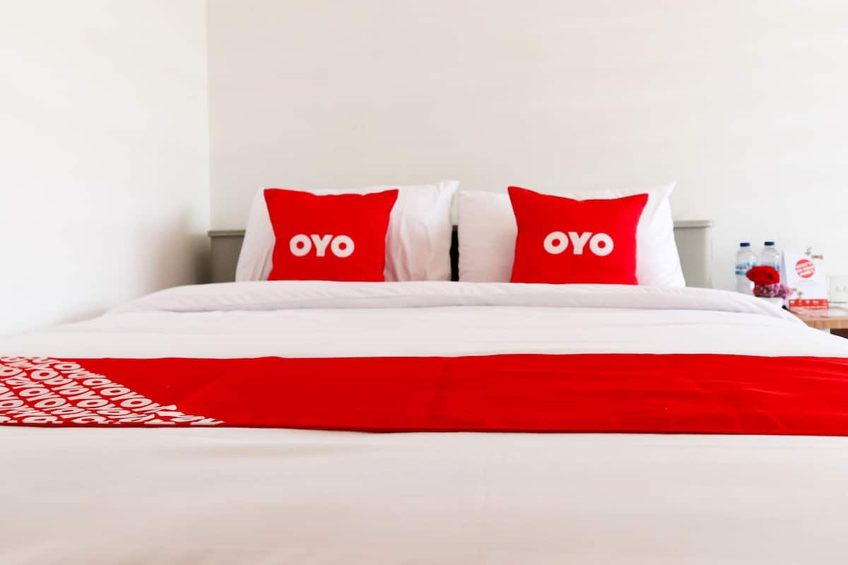A hotel room with a bed, two pillows with the word "OYO" on them, a red and white bedspread, and a nightstand with water bottles and a red flower.