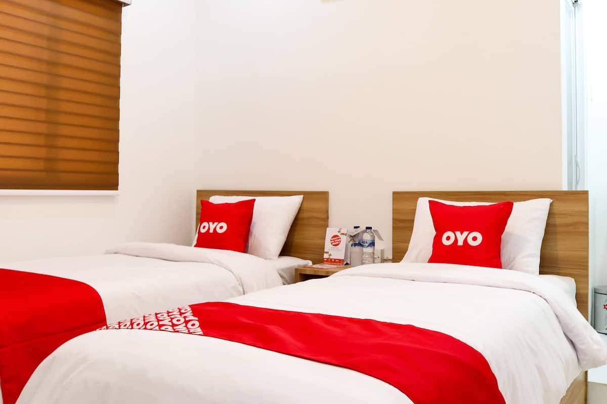 A hotel room with two beds, each with a wooden headboard, white bedding, and red pillows with the word "OYO". There is a nightstand with a water bottle and a small table with a card.