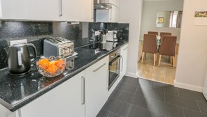 A modern kitchen with a black countertop, a kettle, a toaster, and a coffee maker. There is a dining area with two chairs and a table visible through an open doorway. - Cumbrae View (Largs)