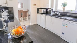 A modern kitchen with white cabinets, a black countertop, and a glass bowl of fruit. There is a dining area with chairs and a table visible through an open door. - Cumbrae View (Largs)
