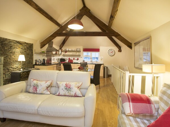 A cozy living room with a white leather sofa, a dining area with a table and chairs, a kitchen in the background, and a stone wall.
